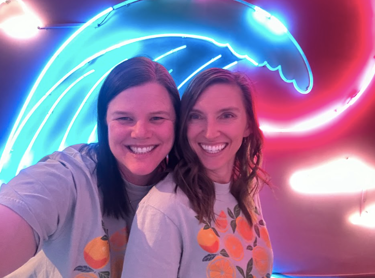 Two women wearing matching t-shirts with images of oranges smile at the camera for a selfie in front of a blue neon wave sign.