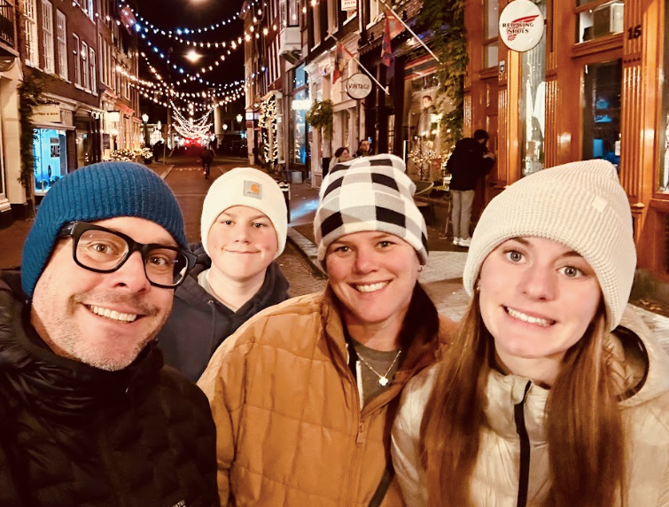 A man in a blue hat and black-rimmed glasses smiles in a selfie with a boy in a white hat, a woman in a black and white checked hat, and a girl in a tan hat.