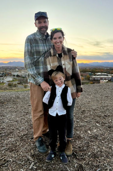 A man in a plaid shirt and a woman in a plaid jacket smile at the camera with a young child dressed up as a cat with black whiskers and ears.