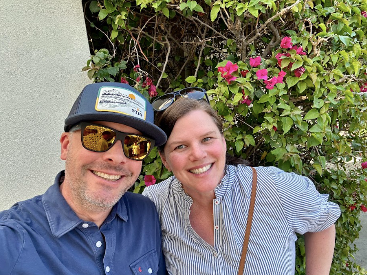 A smiling man in a blue shirt and blue hat smiles at the camera as a woman in a blue and white striped shirt leans in and smiles too.