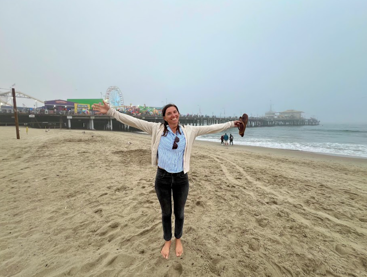 A woman wearing black jeans and a white sweater holds her brown flip-flops in her left hand and holds her arms wide while standing on a beach.