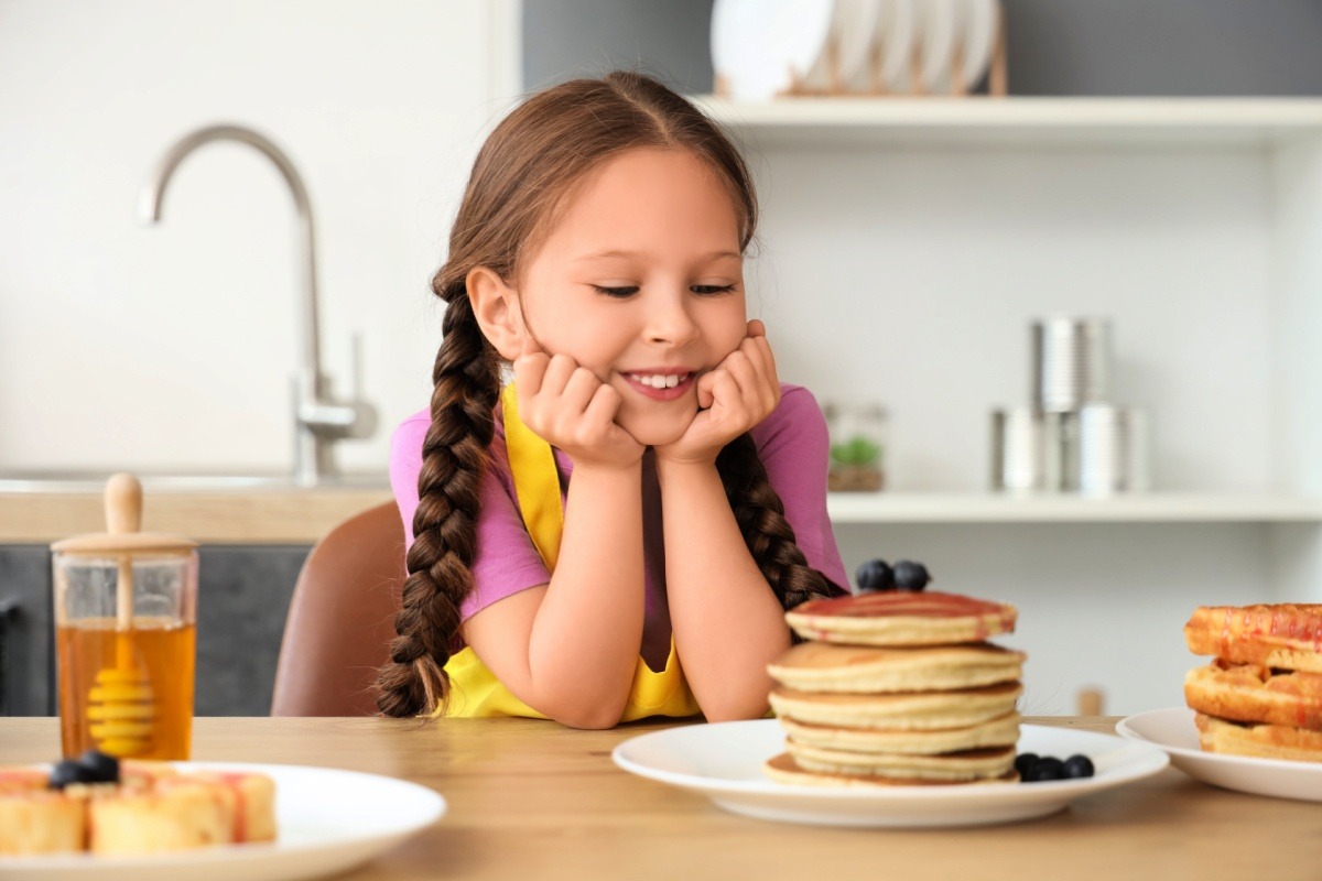 A young girl in a pink t-shirt smiles at a tall stack of pancakes with blueberries on a plate. 