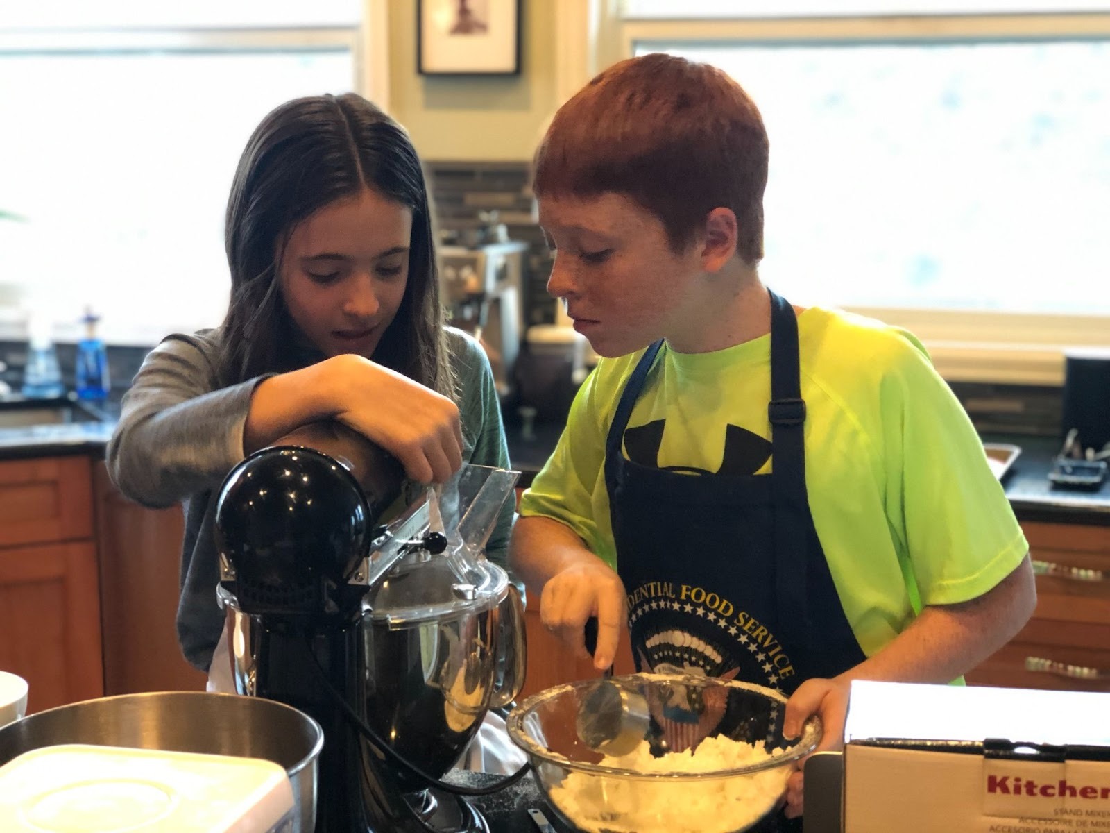 A young girl and a young boy wearing a kitchen apron look closely at a mixing bowl together. 