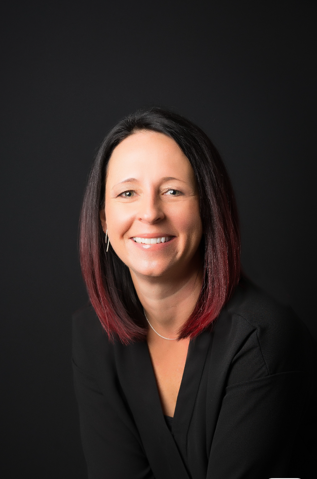 A smiling woman with long dark and red-tipped hair smiles at the camera while wearing a black business suit. 