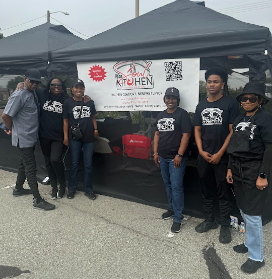 Six people wearing matching black t-shirts stand in front of a booth at a farmer’s market with a sign that reads, “901 Soul Kitchen.”
