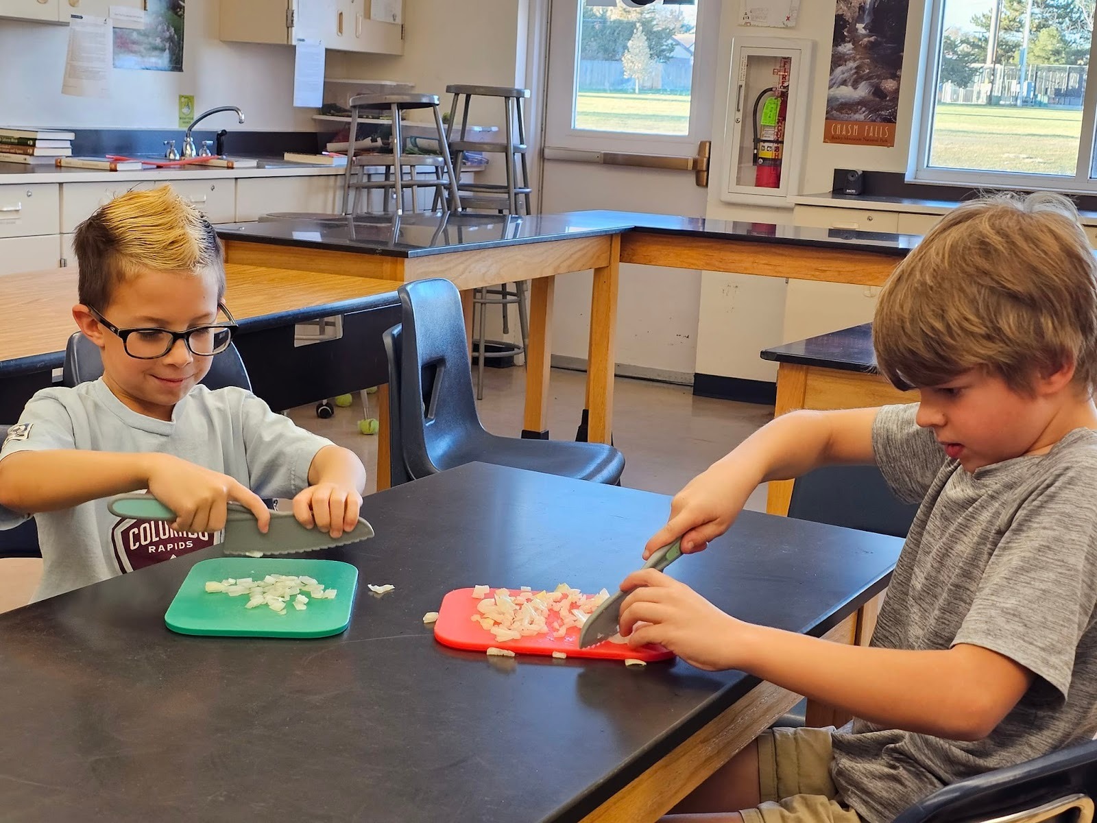 Two young boys sitting at a table in a classroom, use large plastic knives for chopping vegetables during a Sticky Fingers Cooking class.