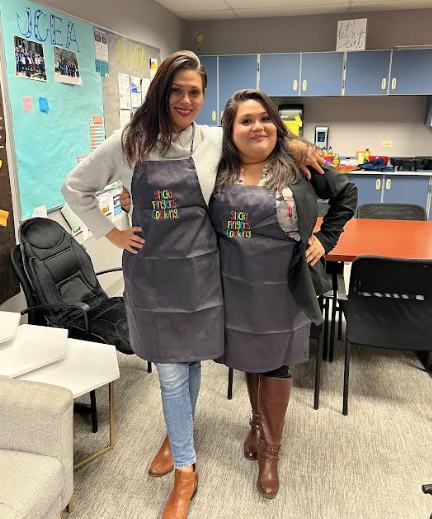 Two women wearing black aprons that read, “Sticky Fingers Cooking” smile at the camera as they stand with their arms wrapped around one another. 