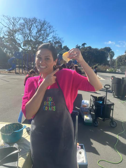 A smiling woman in a pink top with a black apron holds up and points to a piece of food in her hand. 