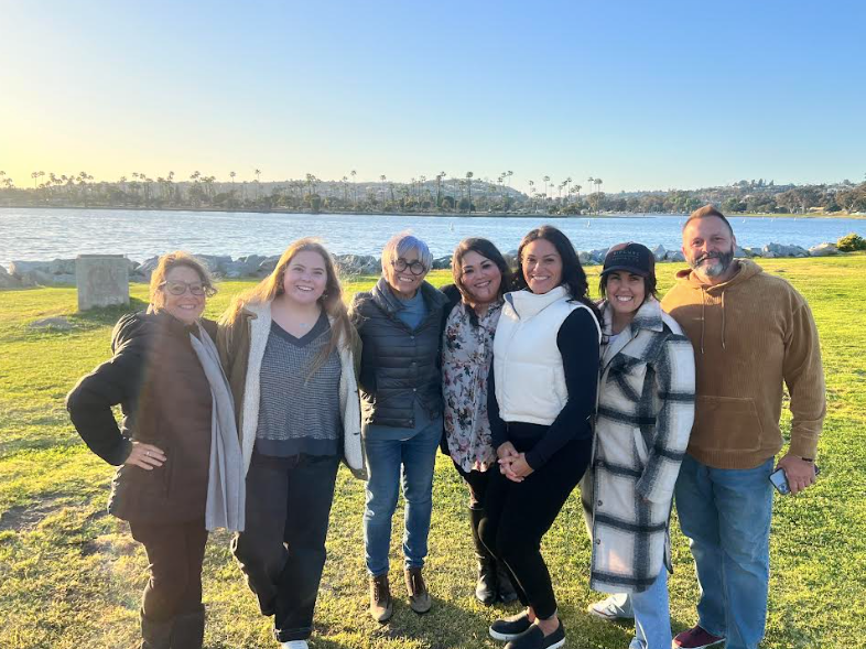 Six women bundled up in warm coats and one man in a brown sweatshirt smile at the camera with a park and lake behind them. 