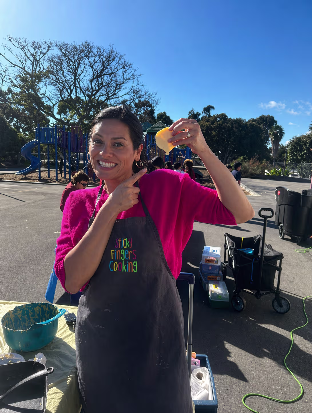 A smiling woman in a pink top with a black apron holds up and points to a piece of food in her hand.