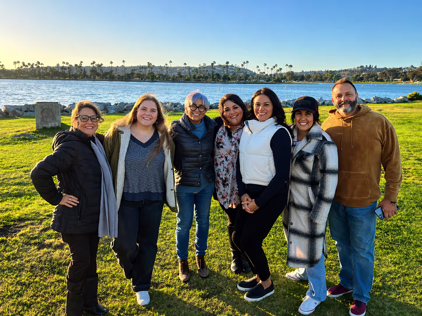 Six women bundled up in warm coats and one man in a brown sweatshirt smile at the camera with a park and lake behind them.