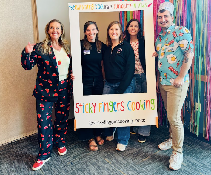 Four smiling women and one smiling man hold up a frame that reads “Sticky Fingers Cooking” in front. 