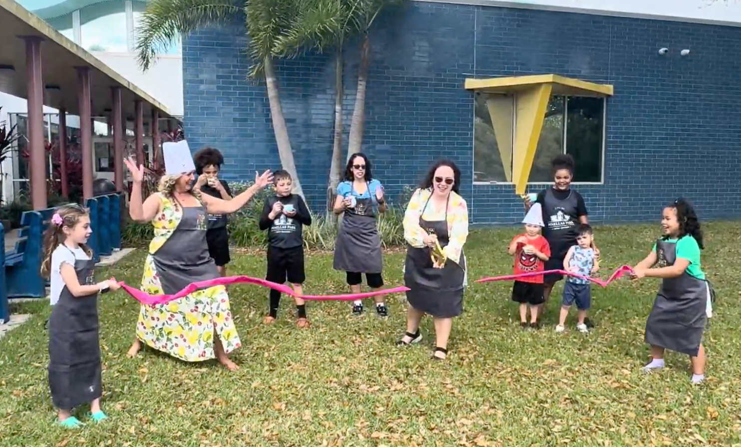 A woman cuts a pink ribbon while children in kitchen aprons cheer her on at the grand opening of Sticky Fingers Cooking of Tampa. 