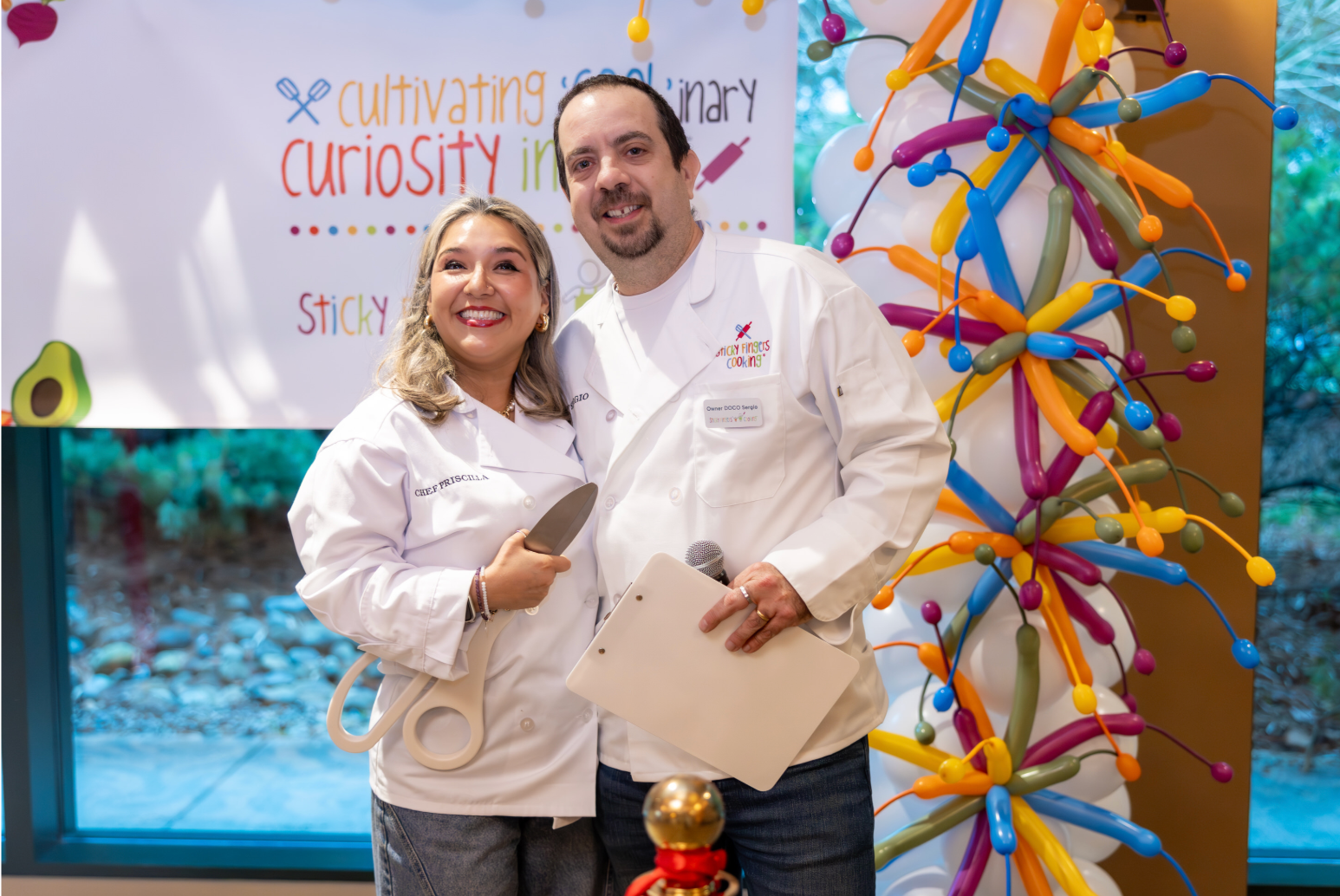 A smiling woman holds a very large pair of scissors and a smiling man holds a microphone at the grand opening of Sticky Fingers Cooking in Seattle East. 
