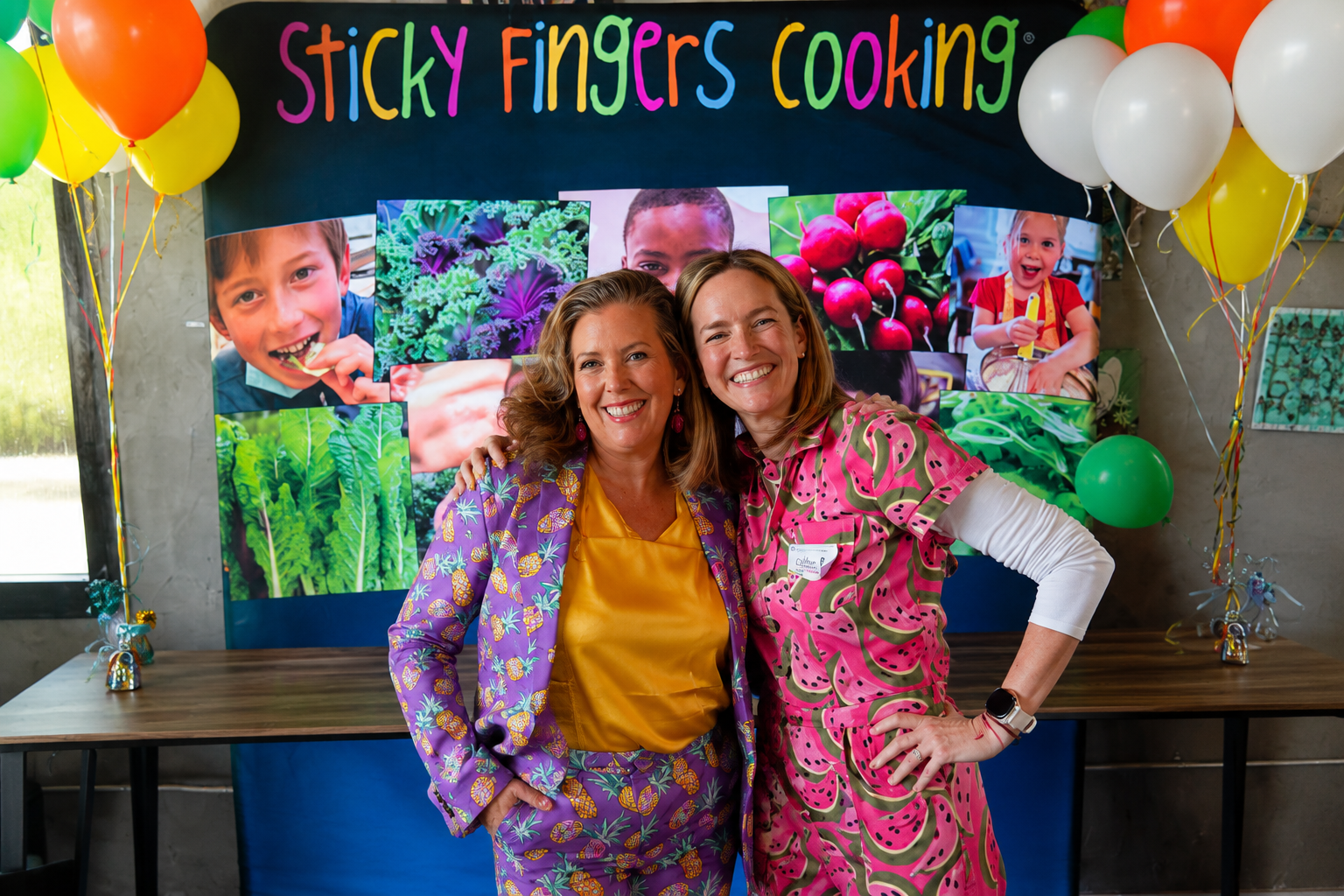 Two women in bright pink and purple colors and surrounded by yellow and white balloons smile at the camera.