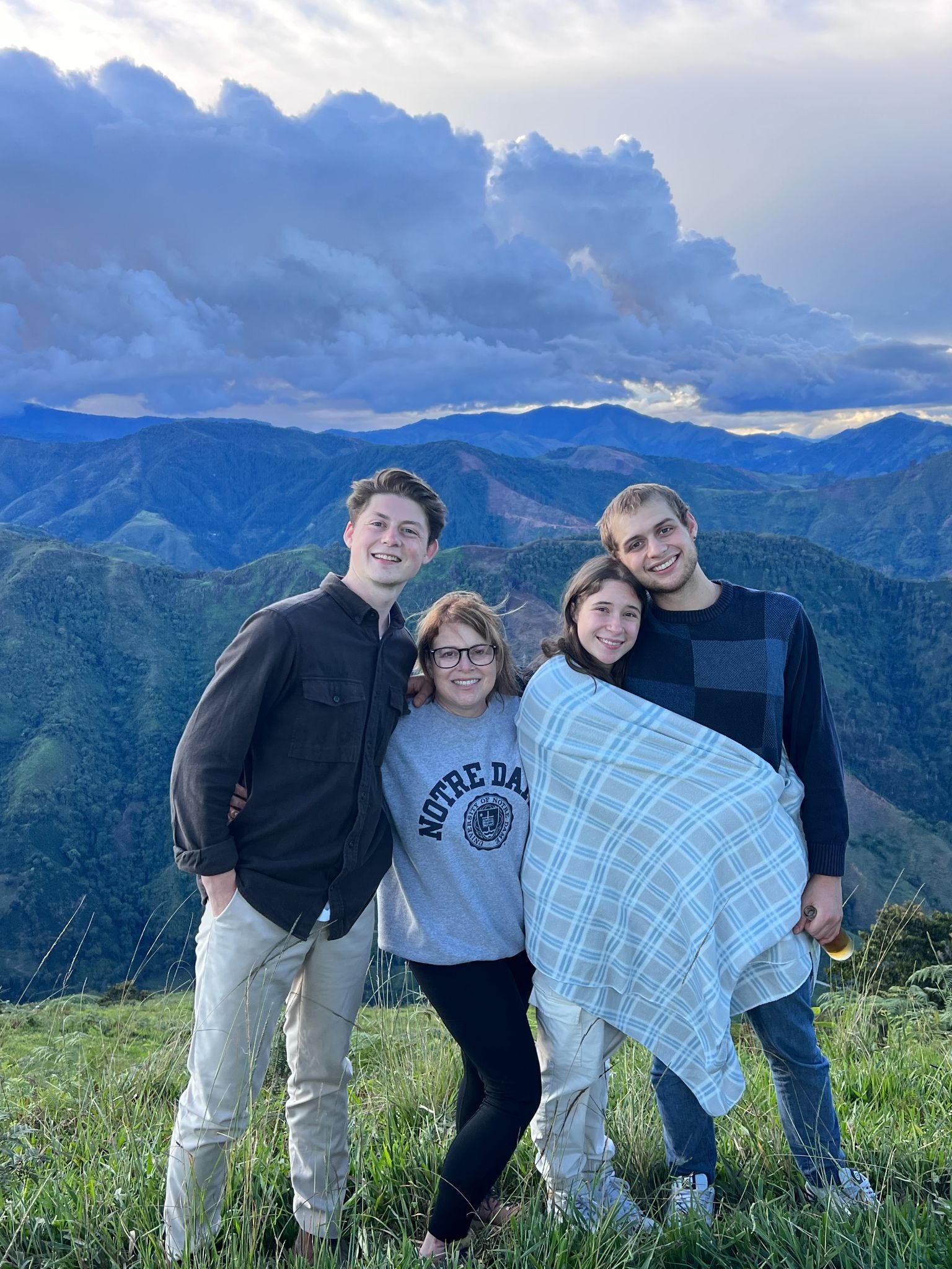 A woman in glasses smiles at the camera with arms wrapped around a teenage daughter and two sons in their 20s with a mountain backdrop. 