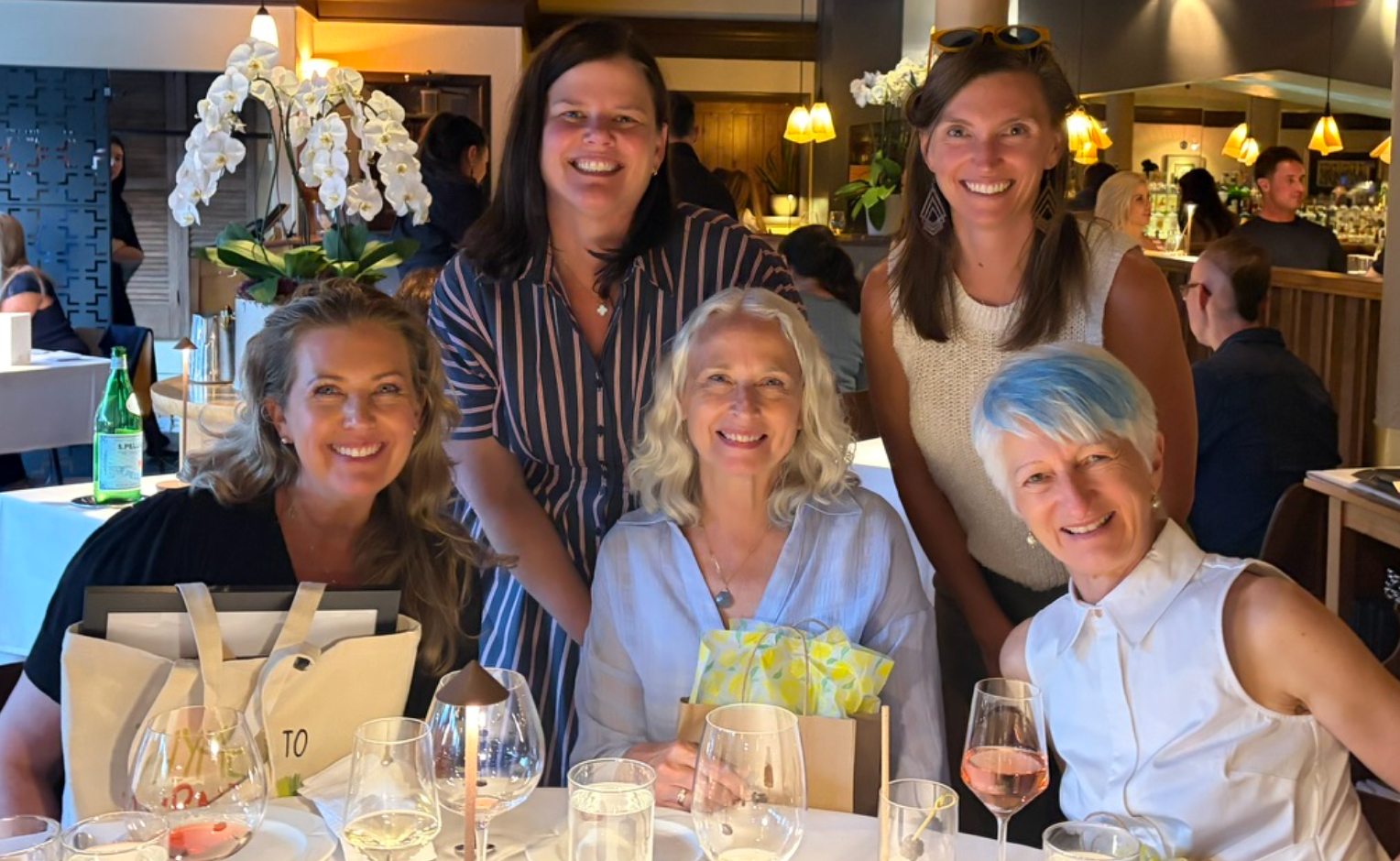 5 women, three seated and two standing behind them, smile at the camera at a nice restaurant