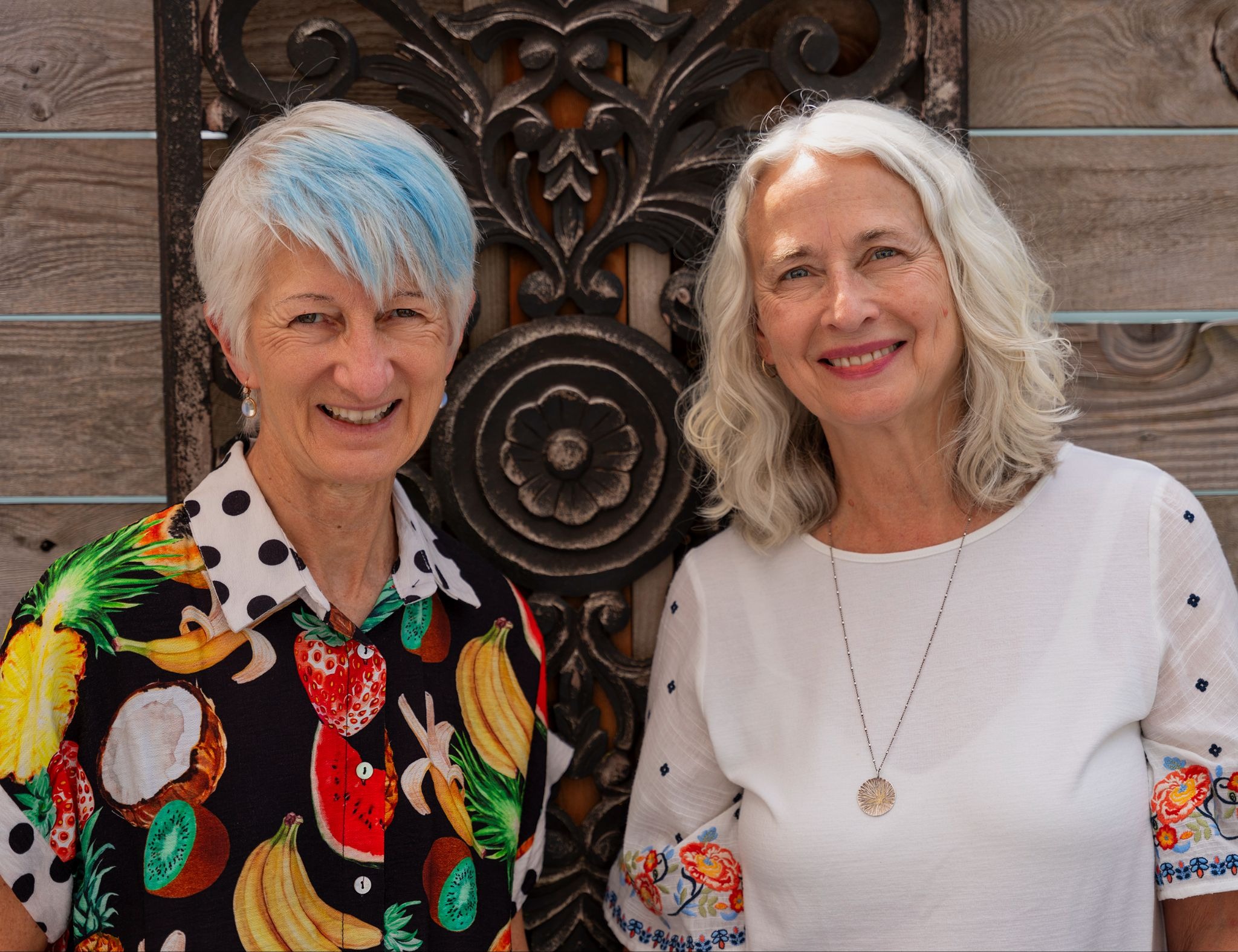 A smiling woman with a blue streak on her white hair and in a colorful shirt with images of fruit stands next to a woman with white hair wearing a white blouse who is also smiling at the camera.
