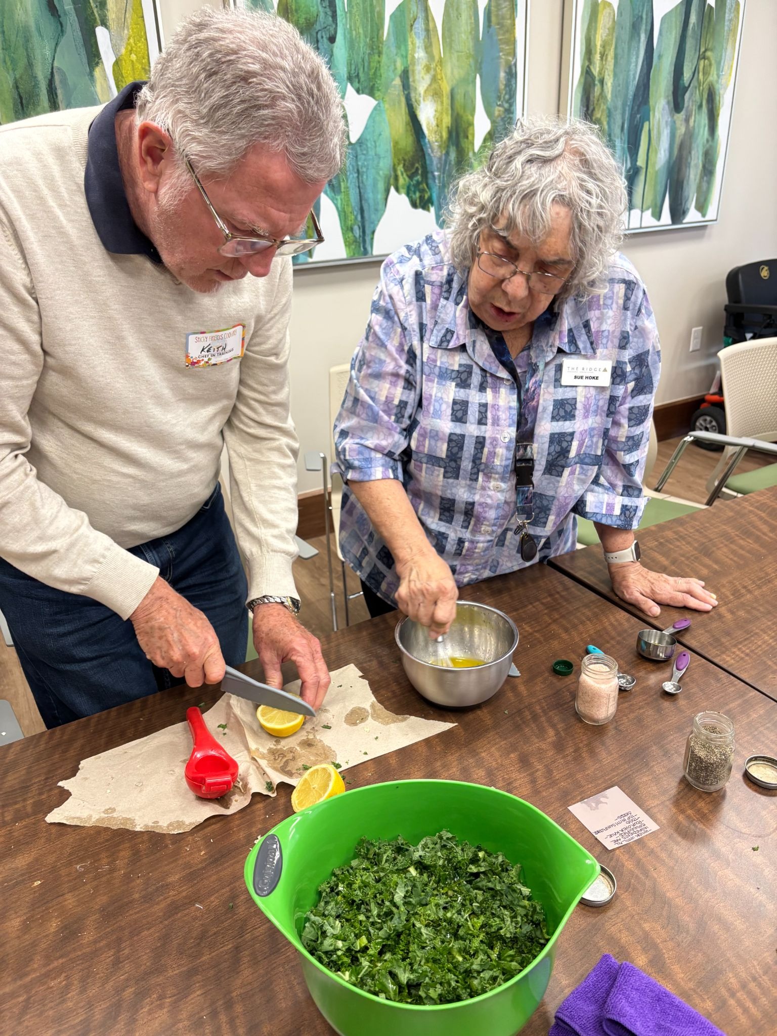 An elderly man slices a lemon as an older woman mixes the juice in a bowl nearby.