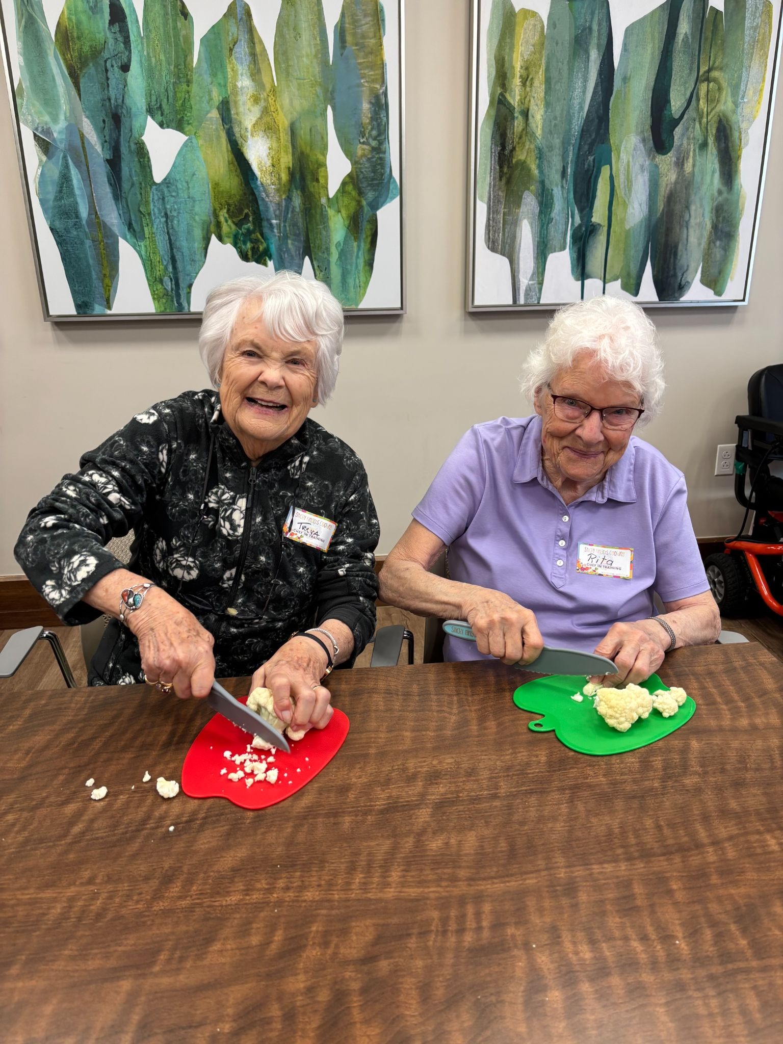Two smiling elderly women chop cauliflower side by side at a table.