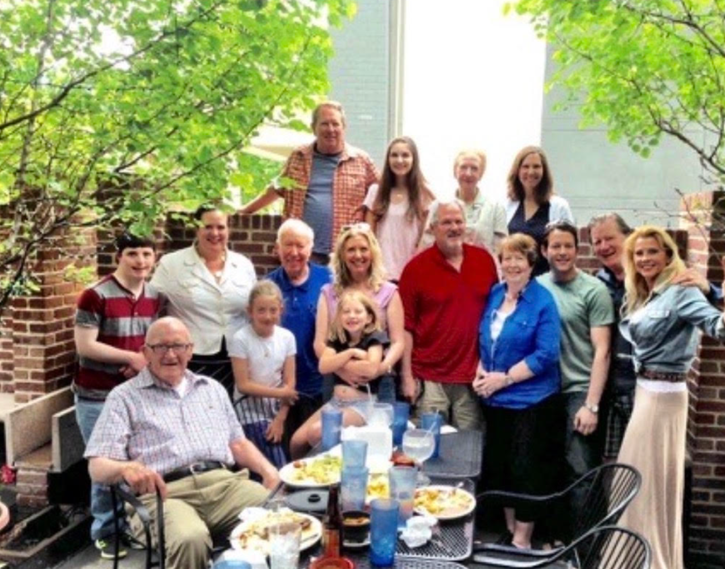 A multi-generational family stands around a table with plates of food as they smile at the camera.