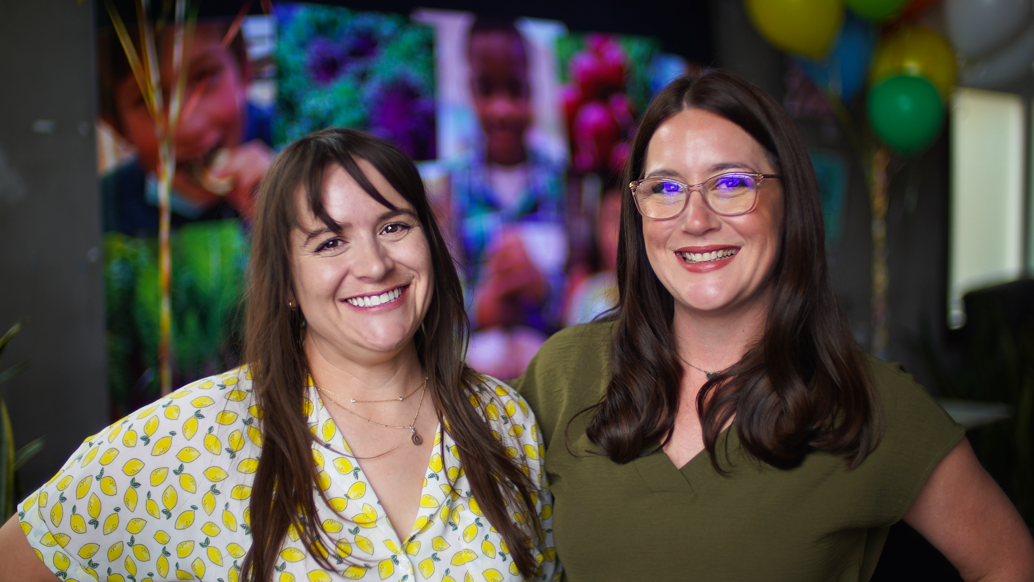 A brunette woman in a shirt with yellow lemons on it and a brunette woman in glasses wearing an olive green top smile at the camera together. 