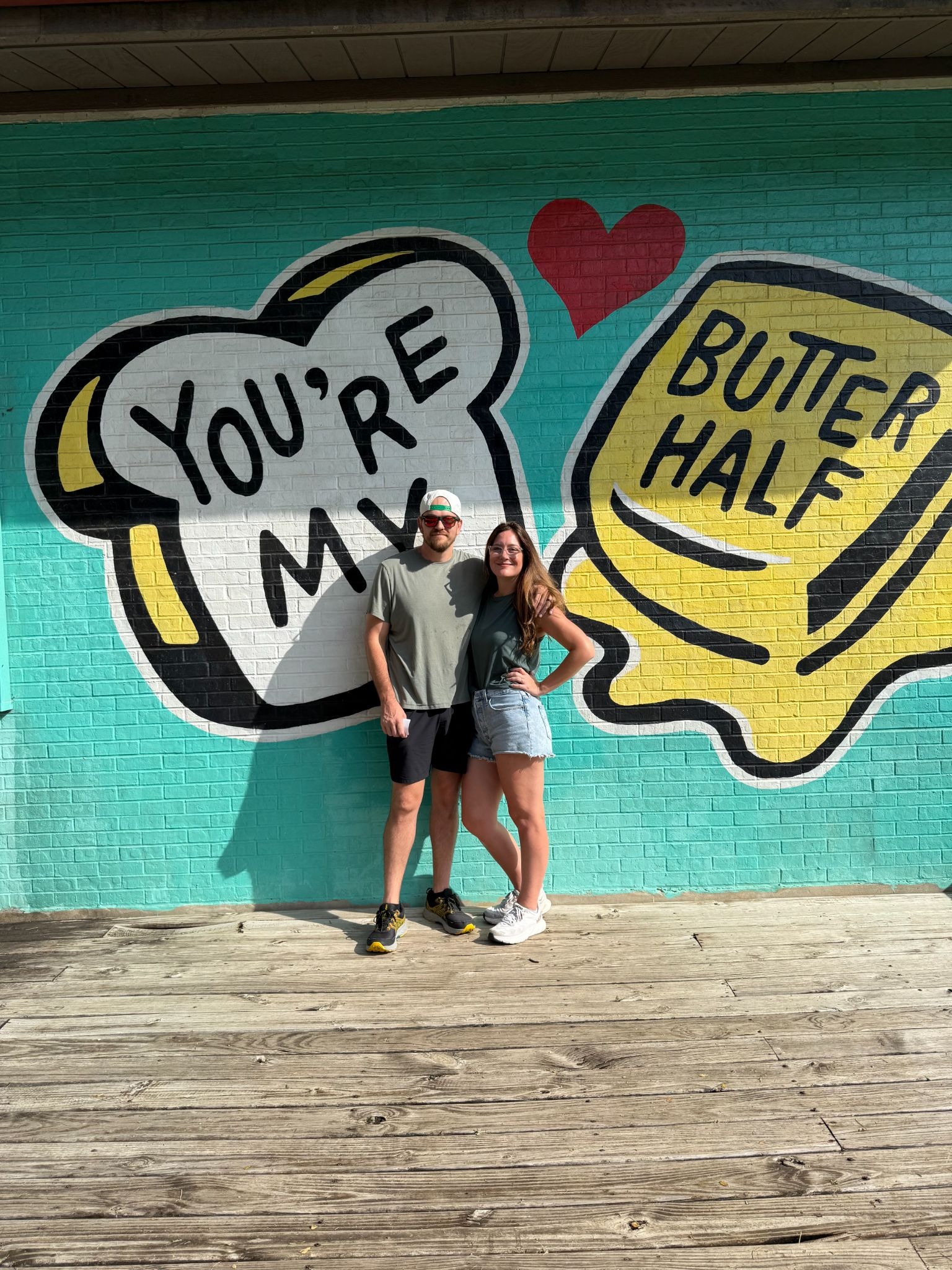 A man in a hat and sunglasses stands with his arm around a woman in cutoff denim shorts and a t-shirt with a sign behind them that reads, ““You’re my BUTTER half!” 