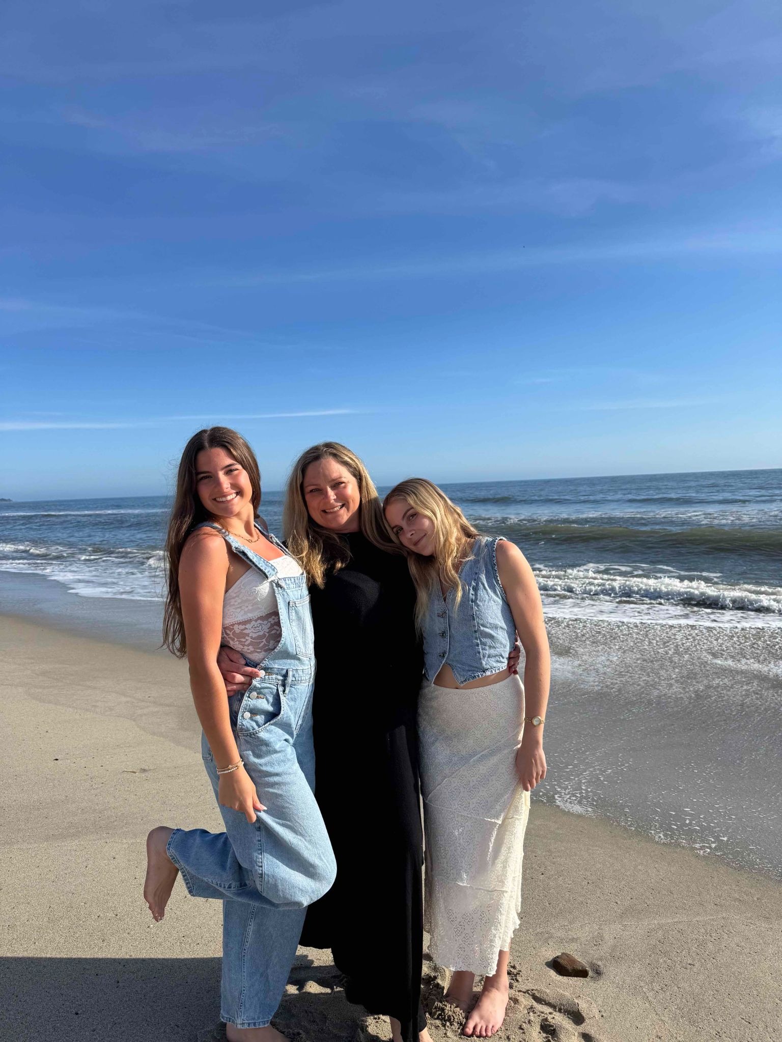 A smiling woman in a long black dress is standing on the beach with two smiling teenage girls.