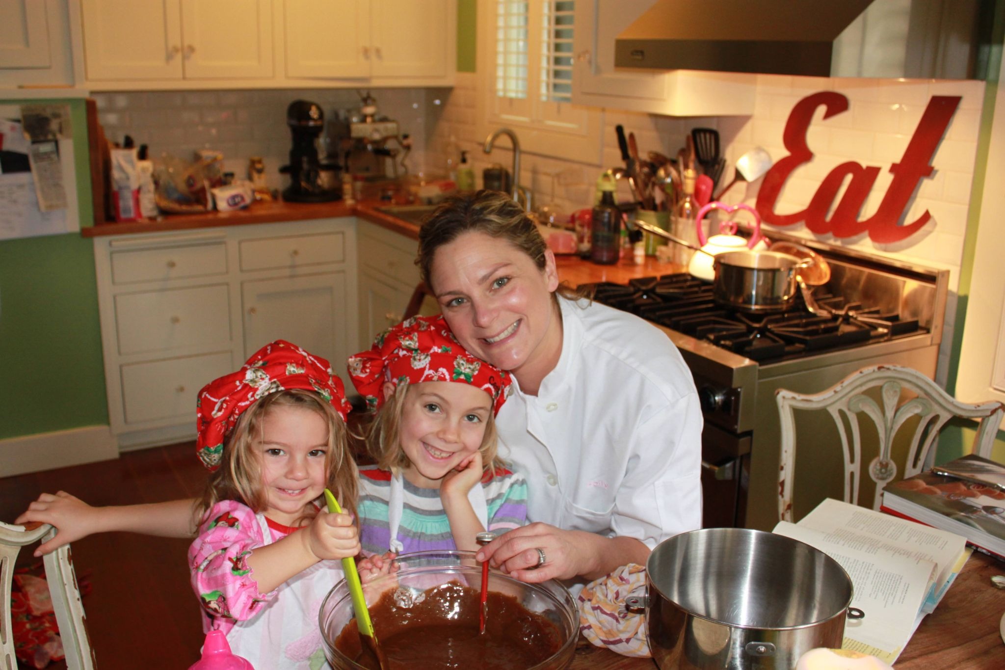 A smiling woman in a white chef coat smiles and leans into two smiling little girls wearing red chef hats while they stir melted chocolate together.