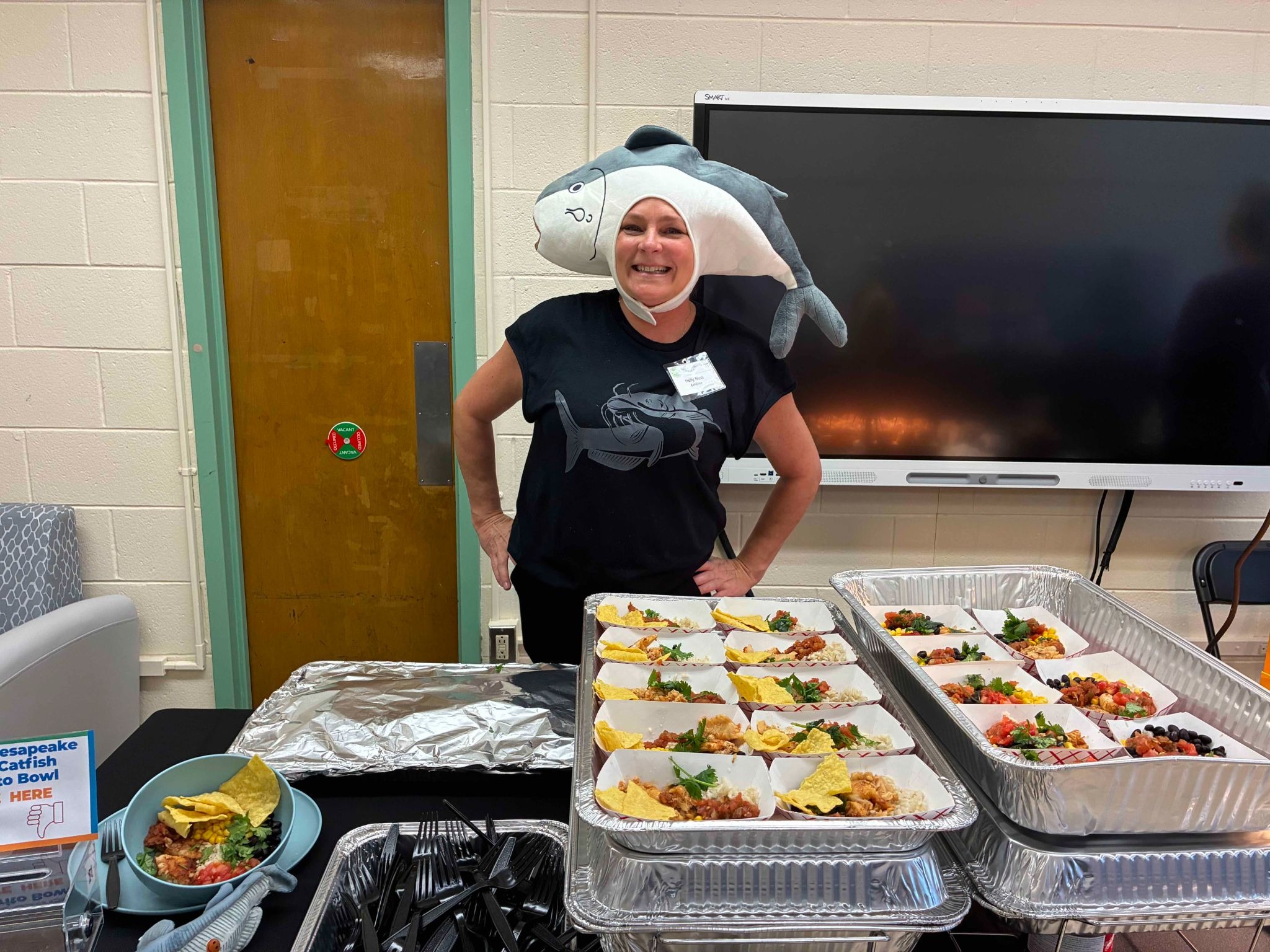 A woman in a black t-shirt grins as she dons a silly shark hat while stanning in a classroom with platters of food.