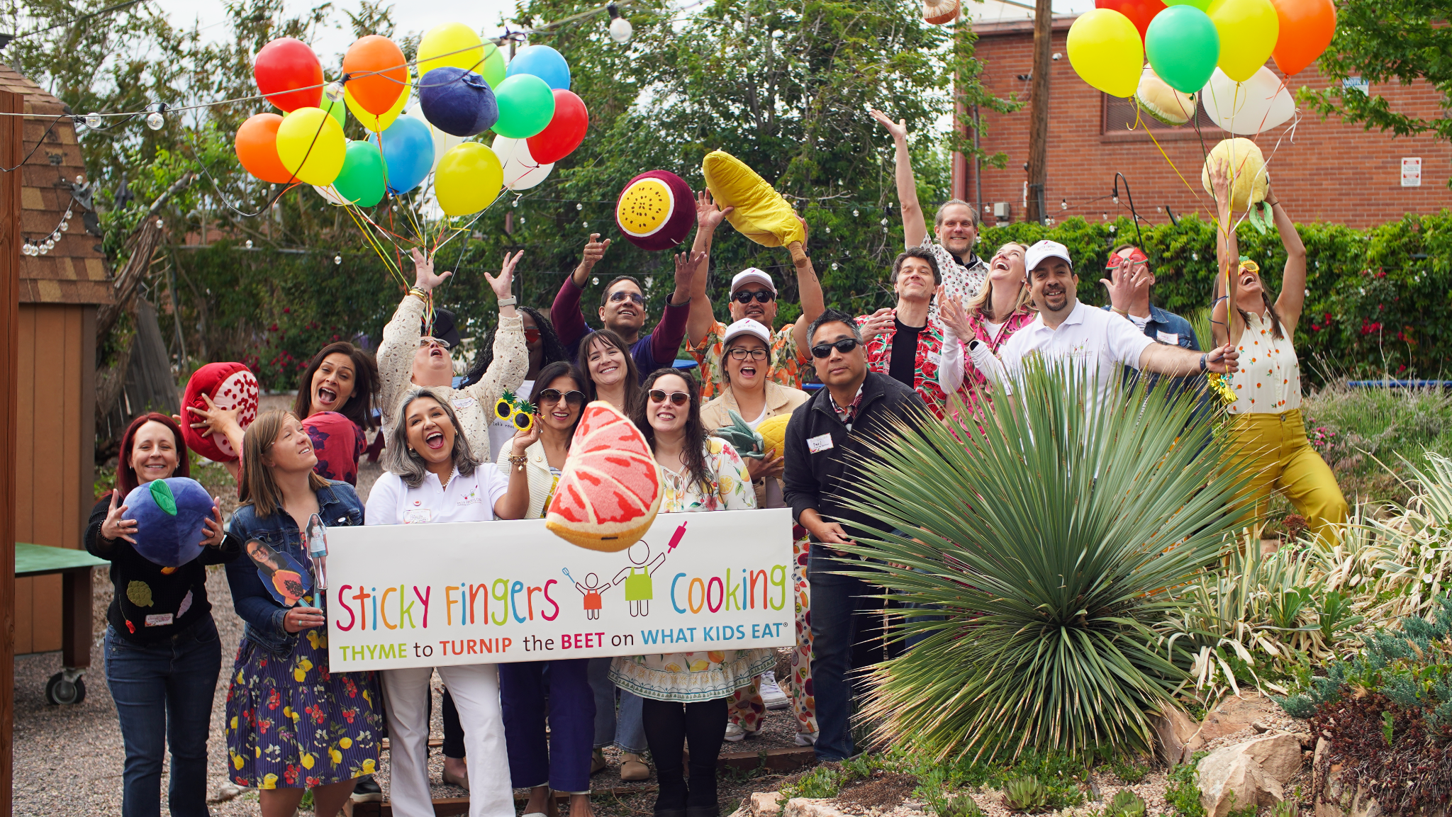 A group of smiling people in sunglasses and tossing pillows that look like fruit gathered together outside.