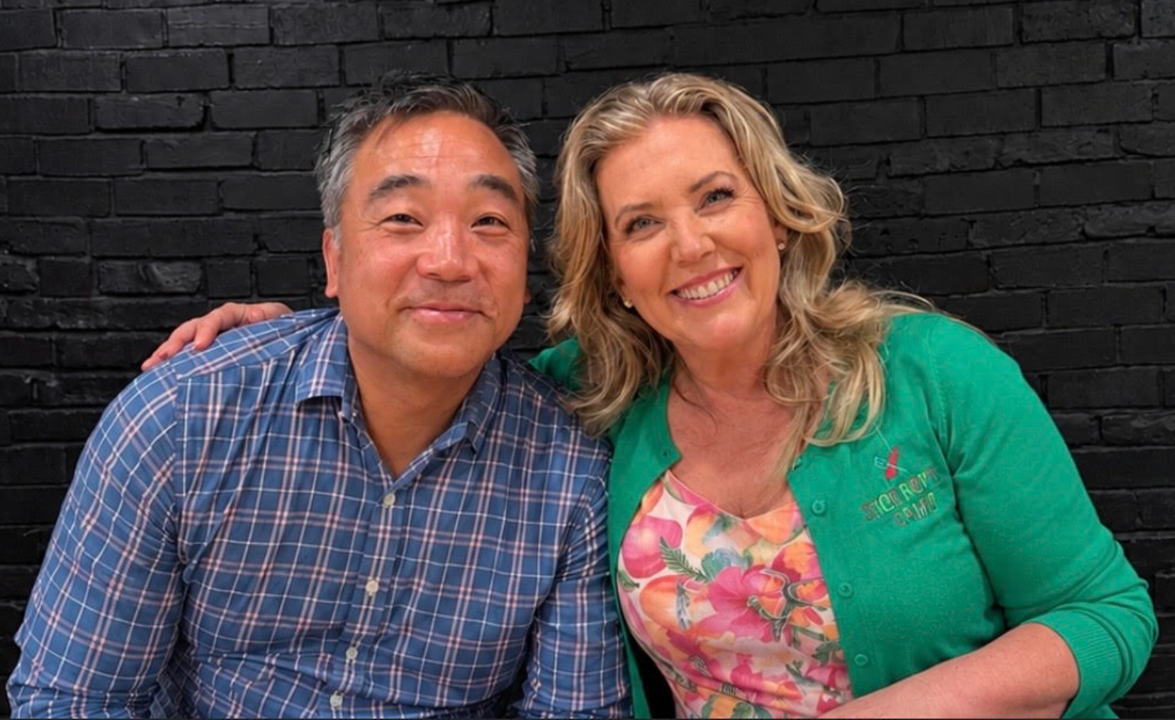A woman in a green sweater puts her arm around a man in a blue plaid shirt as they both smile at the camera.