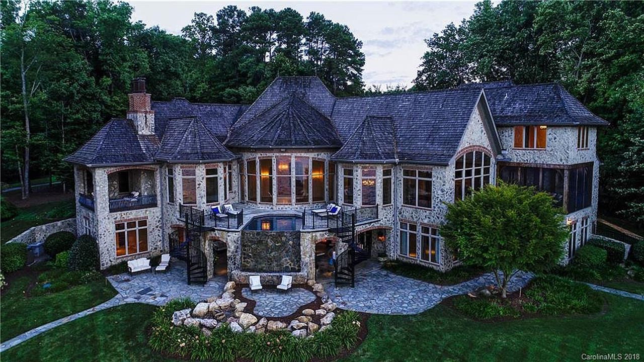 Large stone house with complex roof design, multiple windows with warm interior lighting, outdoor stone patio, spiral staircases, and a landscaped garden with rocks and greenery around the yard.