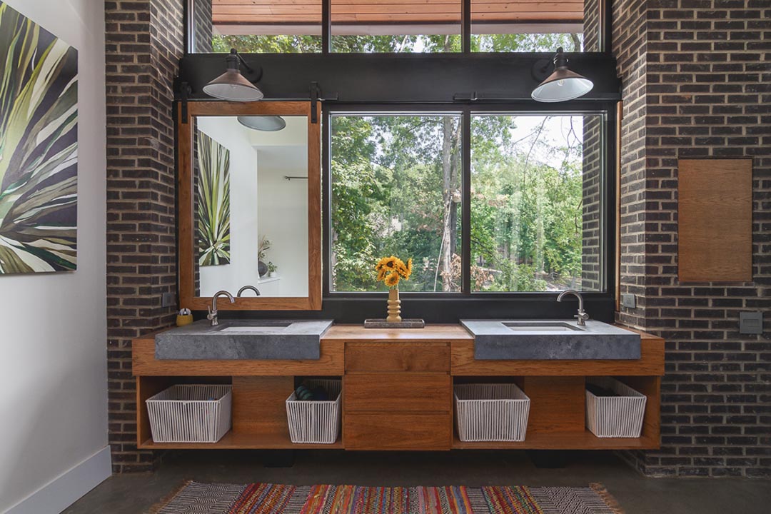 Modern bathroom with a double sink vanity made of wood and concrete basins, a large mirror on the left, a window showing green trees, and a vase with sunflowers on the countertop.