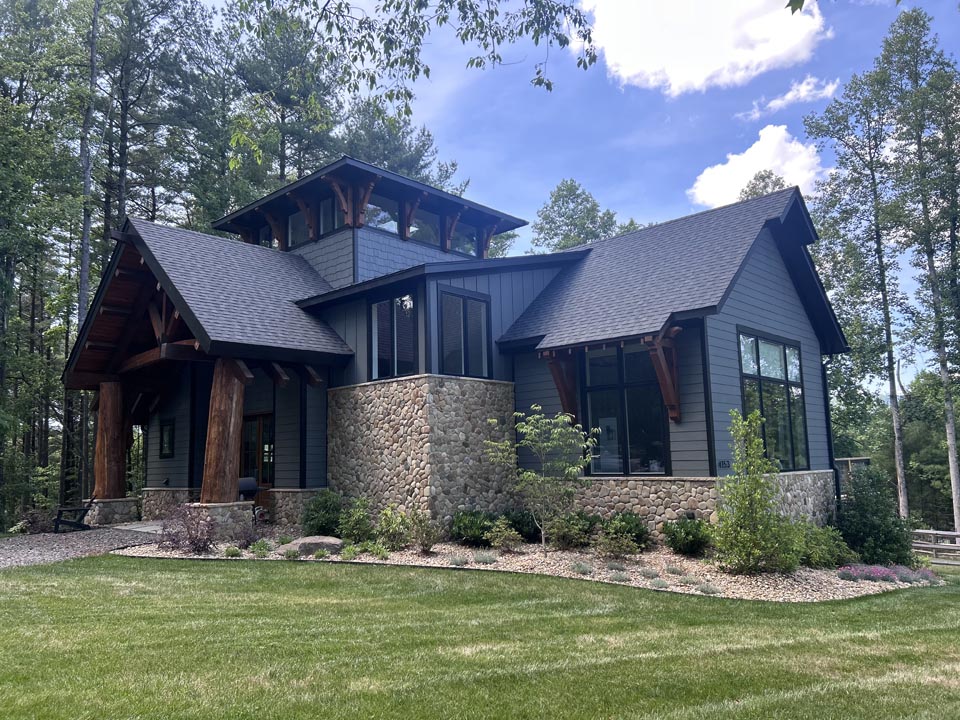 Modern house with dark gray siding, stone foundation, large windows, and wooden columns supporting front porch roof, surrounded by green lawn and tall trees under a partly cloudy blue sky.