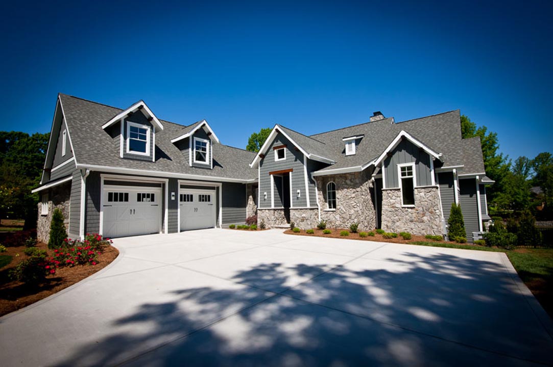 Modern suburban house with gray siding and stone facade, featuring a large concrete driveway and a two-car garage under a clear blue sky.