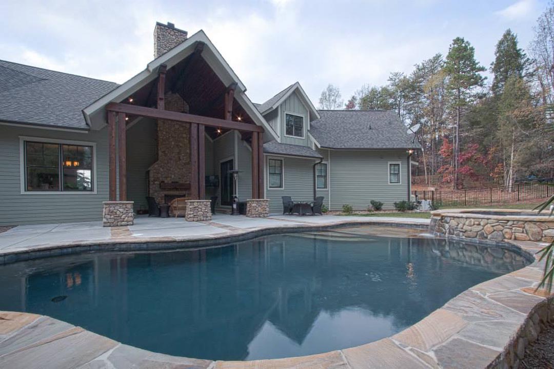 Backyard of a house with a stone-bordered swimming pool in front and a covered patio area supported by wooden beams with stone bases, featuring a stone fireplace, surrounded by trees and lawn.