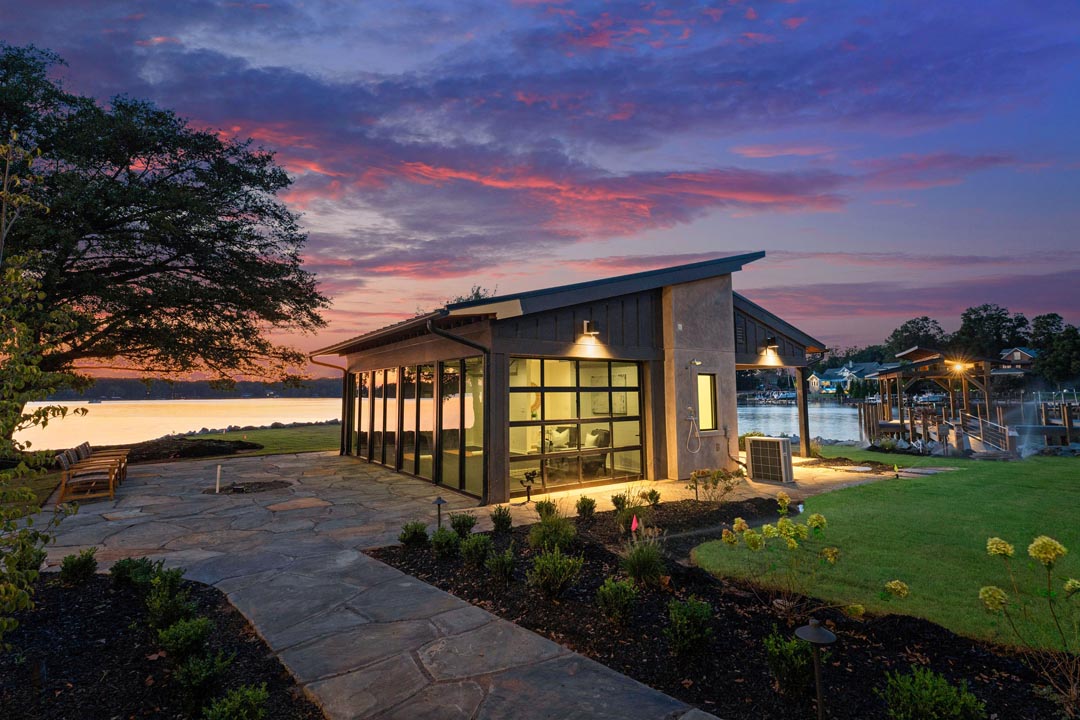 Modern lakeside building with large glass windows at sunset, stone walkway leading to it, lawn, garden beds, and a dock extending into the water.