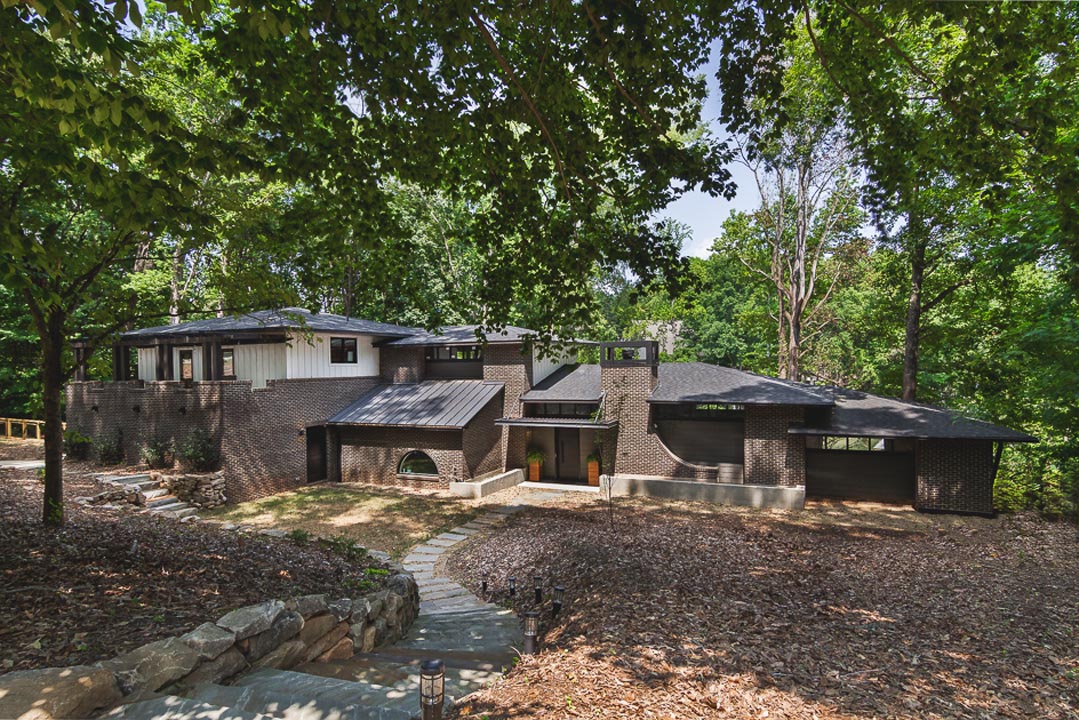 Modern brick house with dark roof surrounded by leafy trees, featuring a curved pathway leading to the entrance and a stone retaining wall along steps.