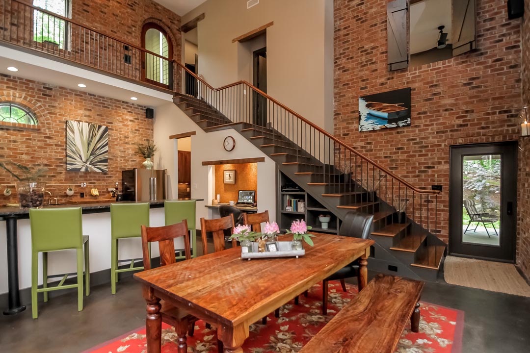 Dining area with a large wooden table, wooden chairs, and a bench on a red floral rug, next to a kitchen bar with three green stools, exposed brick walls, a staircase with wooden treads and black railing, a glass door leading outside, and framed artwork on the walls.