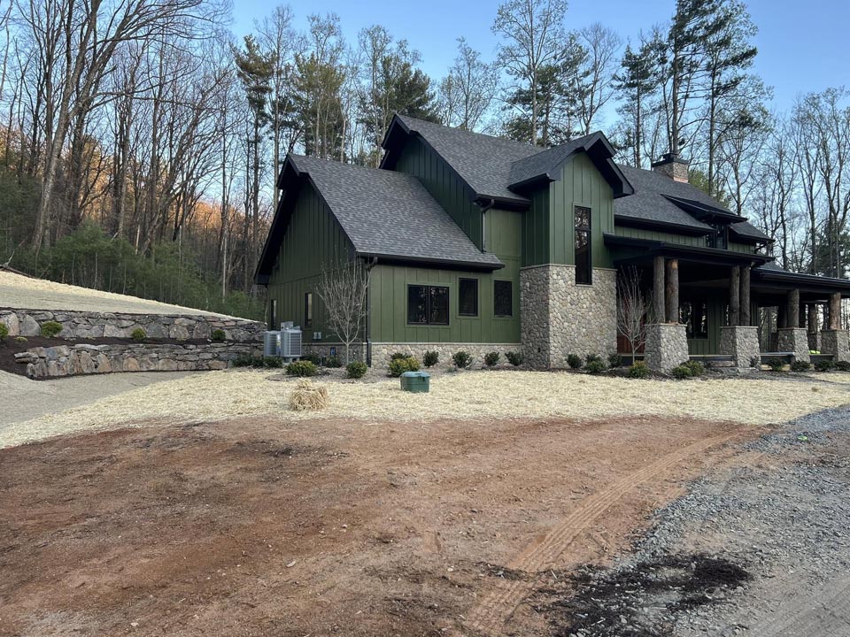 Green and stone two-story house with a covered porch and dark shingled roof, set against leafless trees under a clear blue sky, with a dirt and gravel area in front.