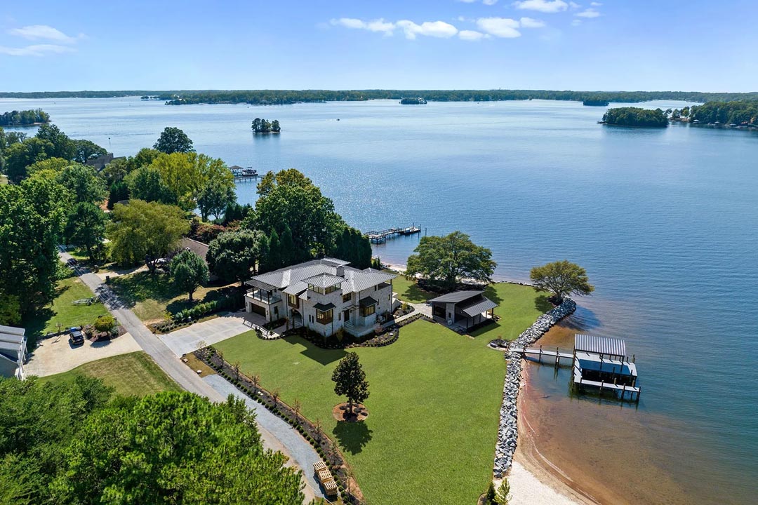 A large house with a gray roof sits on a spacious green lawn by a lake, which has a rocky shoreline, a sandy beach area, a wooden dock with a covered boat lift, and a separate covered pavilion. Trees surround the property, and the lake extends to the horizon with small islands and wooded shorelines visible under a partly cloudy blue sky.