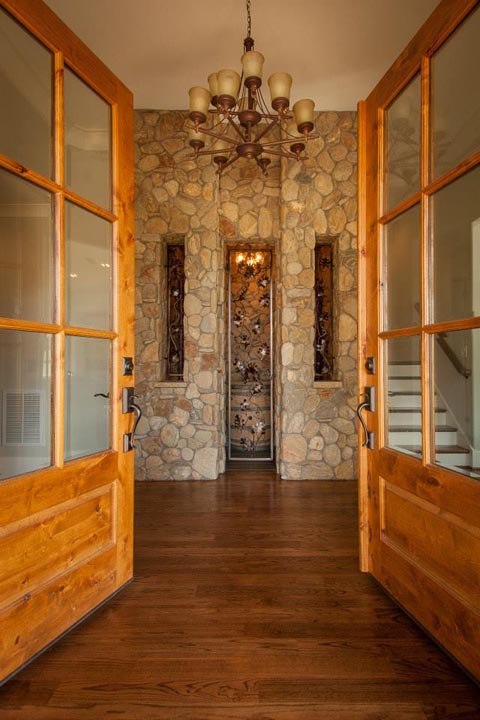 Open wooden double doors leading into a room with a stone wall featuring a decorative metal panel inset and two narrow windows, a wooden floor, and a rustic chandelier hanging from the ceiling.