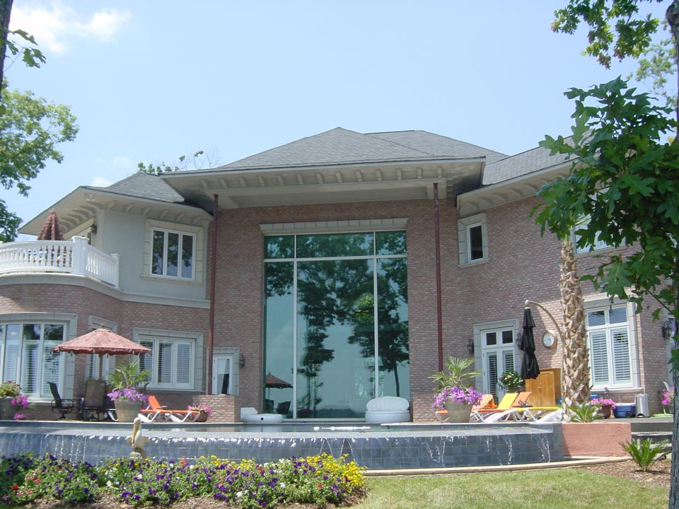 Large two-story brick house with a tall central glass window reflecting trees, surrounded by a patio with lounge chairs, umbrellas, potted plants, and an infinity-style water fountain with cascading water in the foreground and a flower bed with colorful flowers.