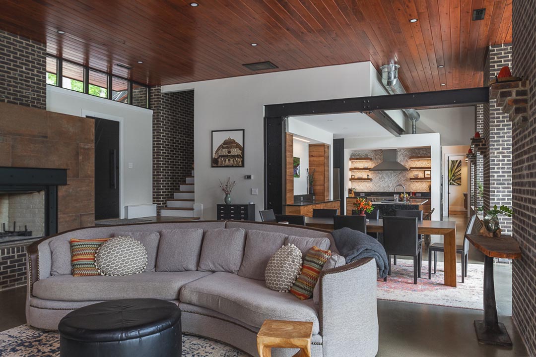 Modern open living space with a curved light gray sofa and patterned pillows centered on a rug, black leather ottoman, wood ceiling, exposed brick walls, fireplace on the left, dining table with black chairs behind the sofa, and a kitchen with wood cabinets and stainless steel appliances in the background.