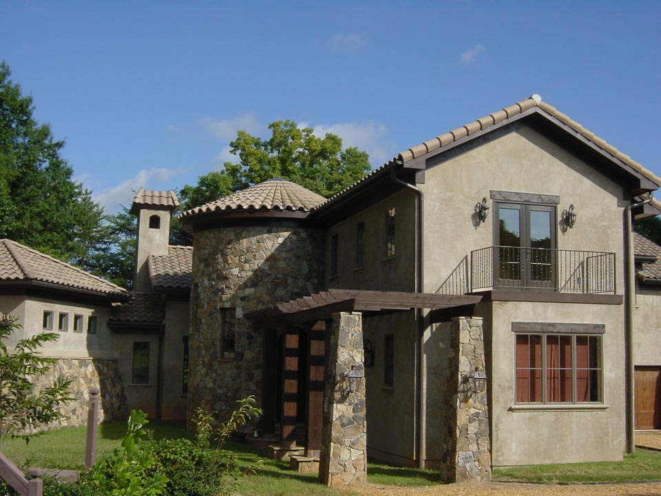 Two-story house with beige stucco and stone facade, round turret with tiled roof, wooden pergola entrance, balcony with metal railing, and a mix of small and large windows under a clear blue sky.