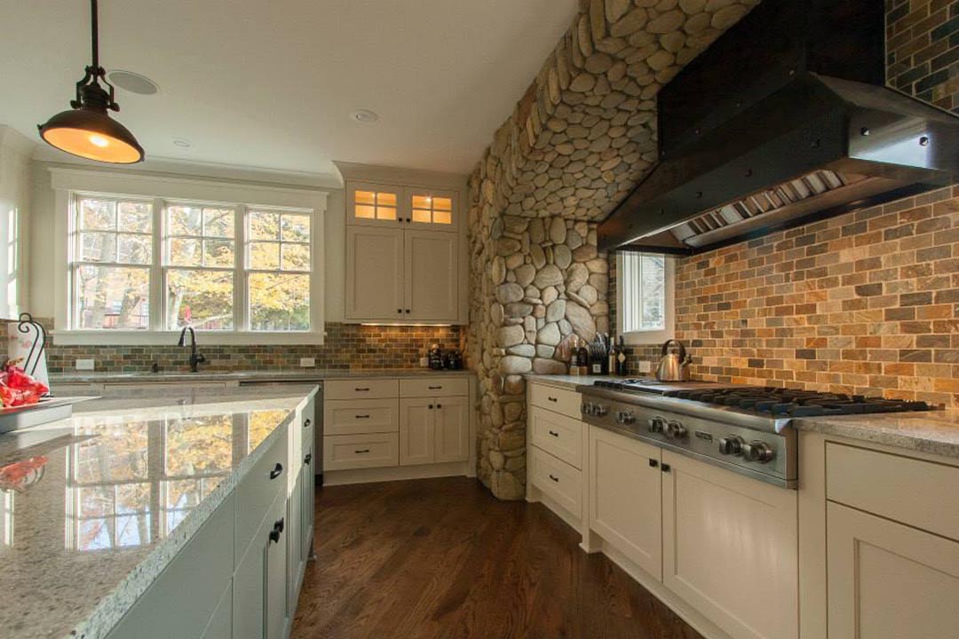 Bright kitchen interior featuring beige cabinets, a large island with granite countertops, a stone accent wall around a stovetop with a stainless steel hood, and a window above the sink letting in natural light, with warm wood flooring and brick-patterned backsplash.
