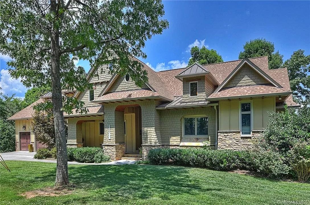 Single-story suburban house with a mix of stone and siding exterior, a brown shingled roof, large front windows, a covered entrance with pillars, and a two-car garage, surrounded by green grass, bushes, and trees under a blue sky with scattered clouds.