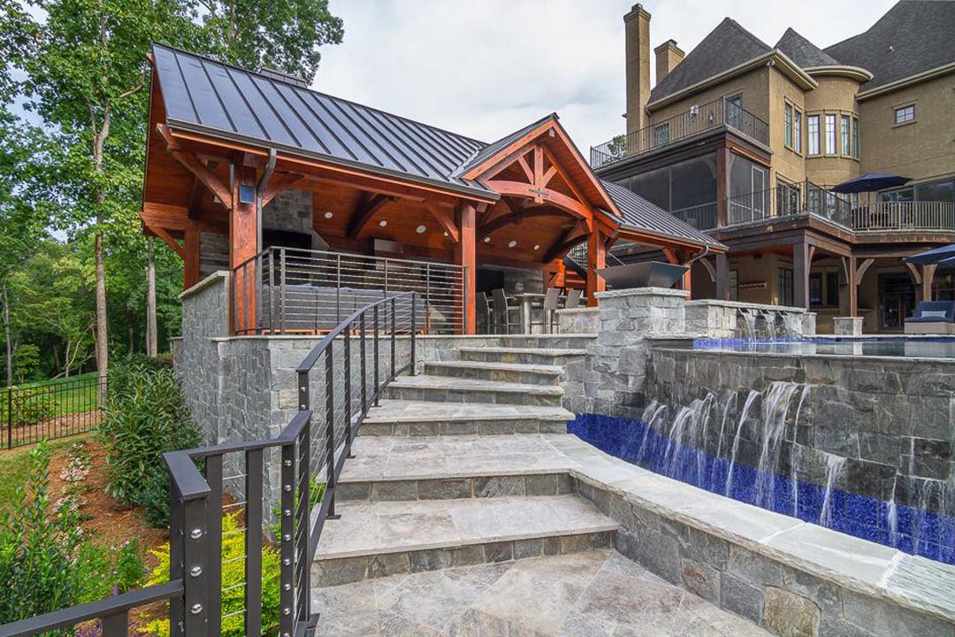 Stone stairs with metal railing leading up to a covered patio with wooden beams and metal roof next to a cascading water waterfall feature, surrounded by a large house and green trees.
