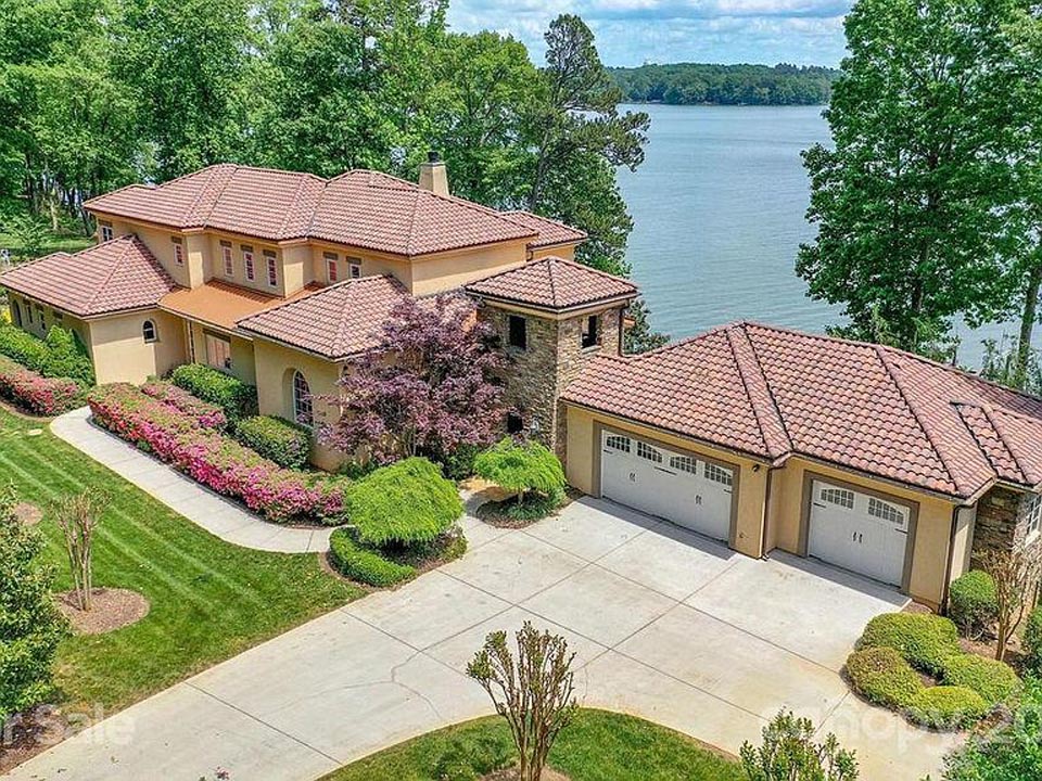 Large two-story house with a terracotta tile roof and beige walls, surrounded by green trees, colorful flowering bushes, and a manicured lawn, located near a calm lake in the background. The driveway leads to a three-car garage attached to the house.